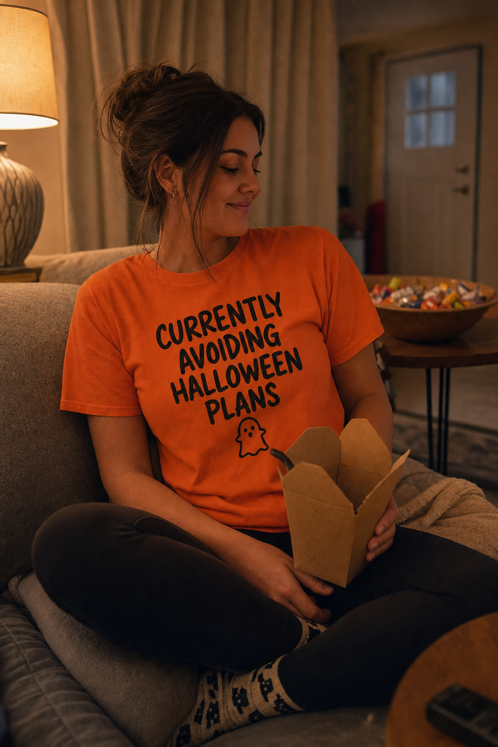 Woman on a sofa at home on Halloween night wearing the orange Currently Avoiding Halloween Plans tee
