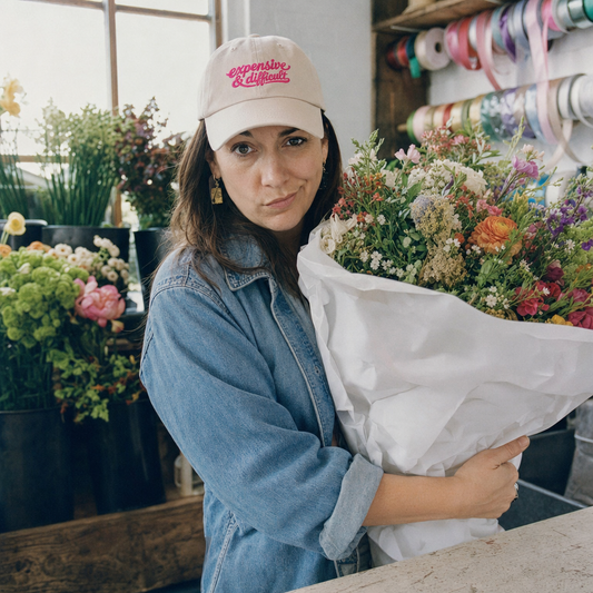 Stone classic cap embroidered “expensive & difficult” in pink script, worn at a florist’s counter